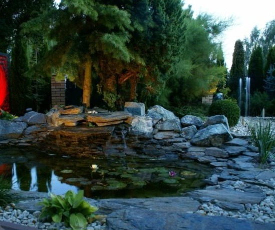 Oczko wodne z kamienną kaskadą, otoczone zielenią i oświetlone punktowo, z fontanną w tle. Elementy dekoracyjne z kamienia i roślin wodnych.