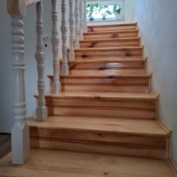 Andrzej Werner uslugi remontowo budowlane - Jasne, drewniane schody z białymi balustradami prowadzą w górę, oświetlone światłem z okna z roślinami. Widok z dołu, ukazujący detale drewna i wykończenia.