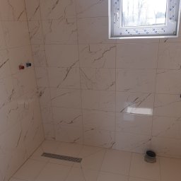 Andrzej Werner uslugi remontowo budowlane - Wykończona łazienka z beżowymi płytkami imitującymi marmur, widoczne okno, odpływ liniowy i przyłącza wodne. Minimalistyczny design.