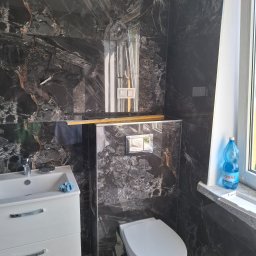 Andrzej Werner uslugi remontowo budowlane - Elegancka łazienka po remoncie: czarne płytki imitujące marmur na ścianach, biała umywalka z szafką i toaleta. Butelka wody na parapecie.