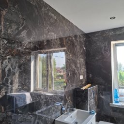 Andrzej Werner uslugi remontowo budowlane - Elegancka łazienka z czarnymi płytkami imitującymi marmur, biała umywalka z szafką i toaleta. Okno z widokiem na zieleń. Nowoczesne oświetlenie sufitowe.