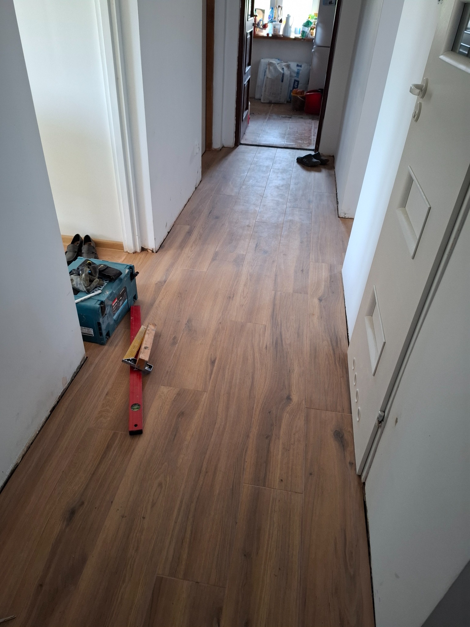 Układanie paneli podłogowych w korytarzu, widoczna skrzynka z narzędziami, poziomica i docięte panele. Częściowo widoczne drzwi i wejście do innego pomieszczenia.