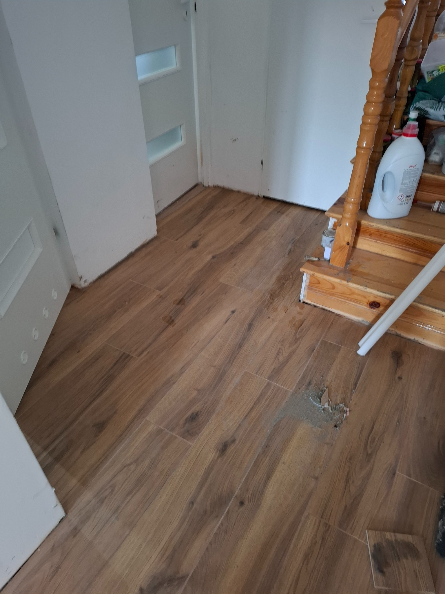 Ułożona podłoga z płytek imitujących drewno w korytarzu, widoczne schody z drewnianą balustradą i białe drzwi z przeszkleniami. Fragment uszkodzonej podłogi.
