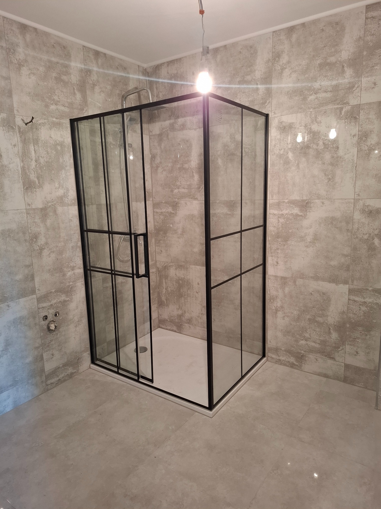 Nowoczesna kabina prysznicowa z czarnymi profilami i szklanymi ściankami, zamontowana w łazience wykończonej płytkami imitującymi beton. Widoczne przyłącza wodne.