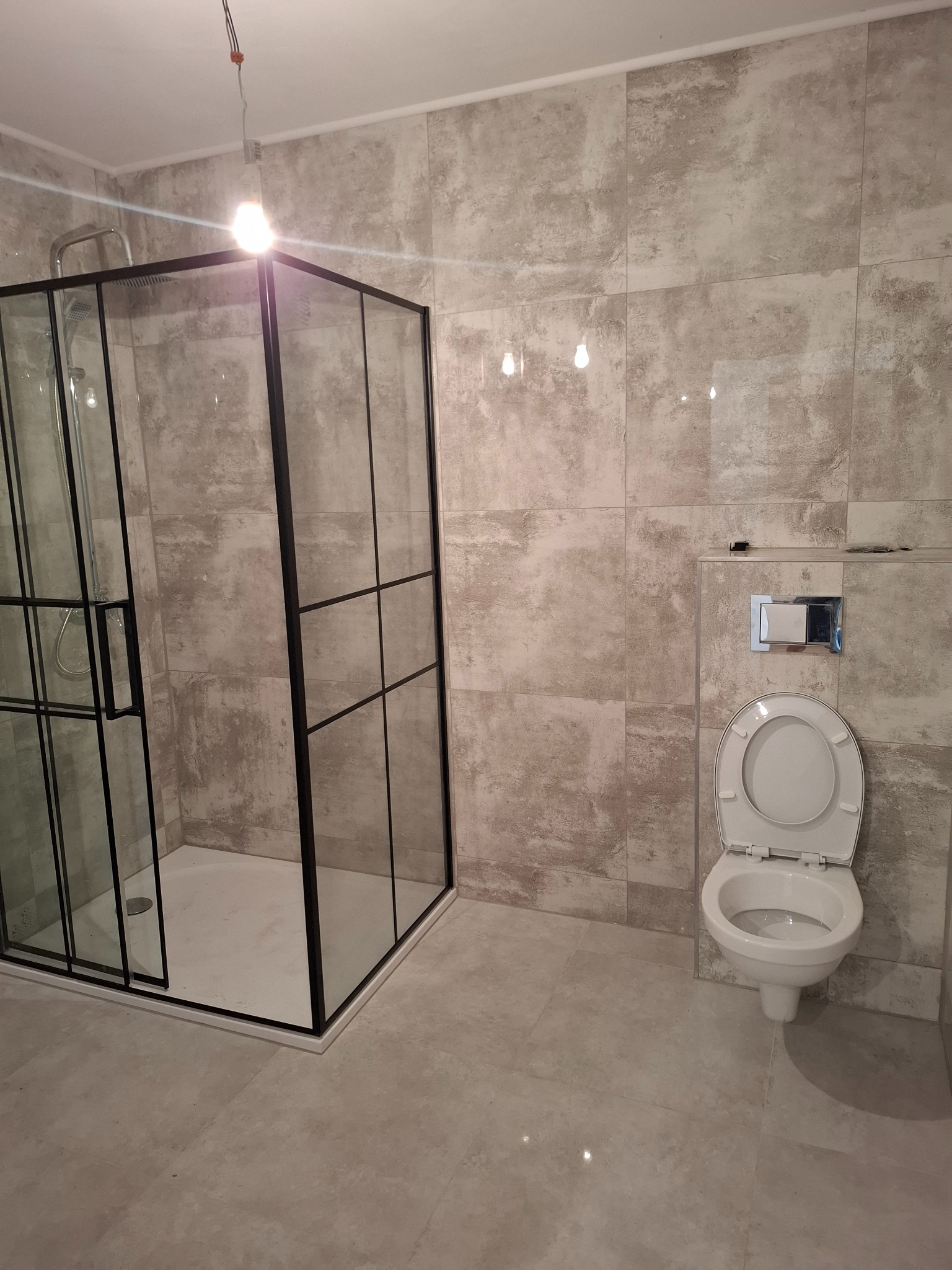 Wykończona łazienka z kabiną prysznicową typu walk-in z czarnymi profilami, toaletą i płytkami imitującymi kamień na ścianach i podłodze. Widoczne niedokończone podłączenie oświetlenia.