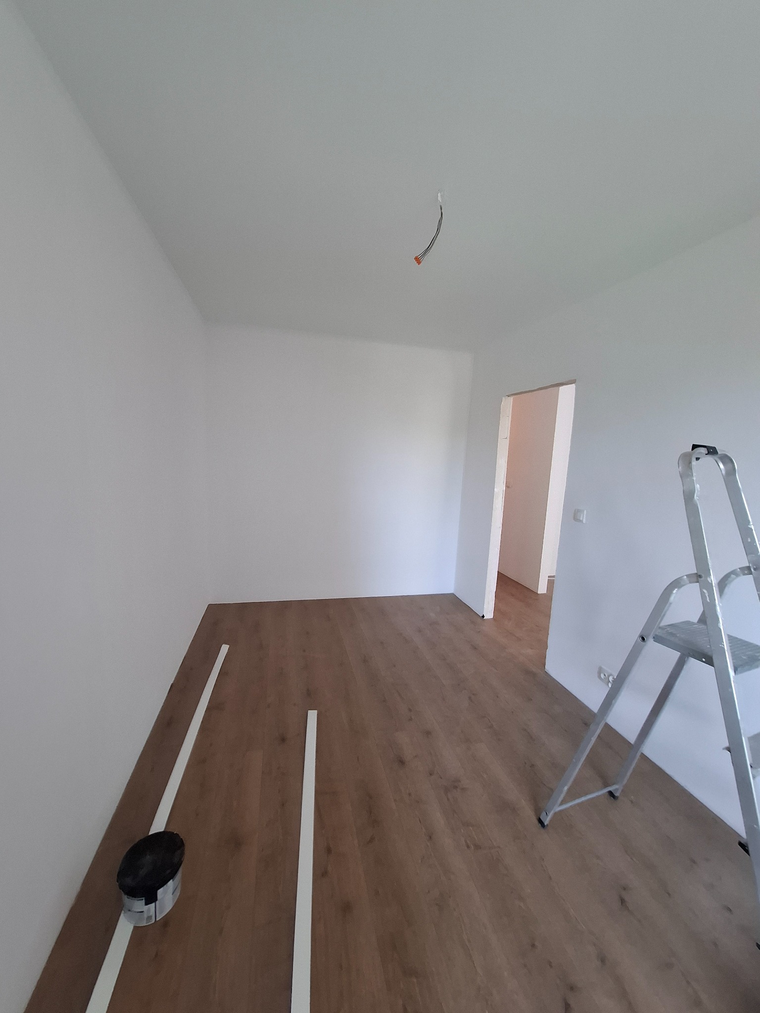 Pokój z panelami podłogowymi w trakcie montażu, widoczna drabina i listwy przypodłogowe. W tle białe ściany i otwarte drzwi do innego pomieszczenia.