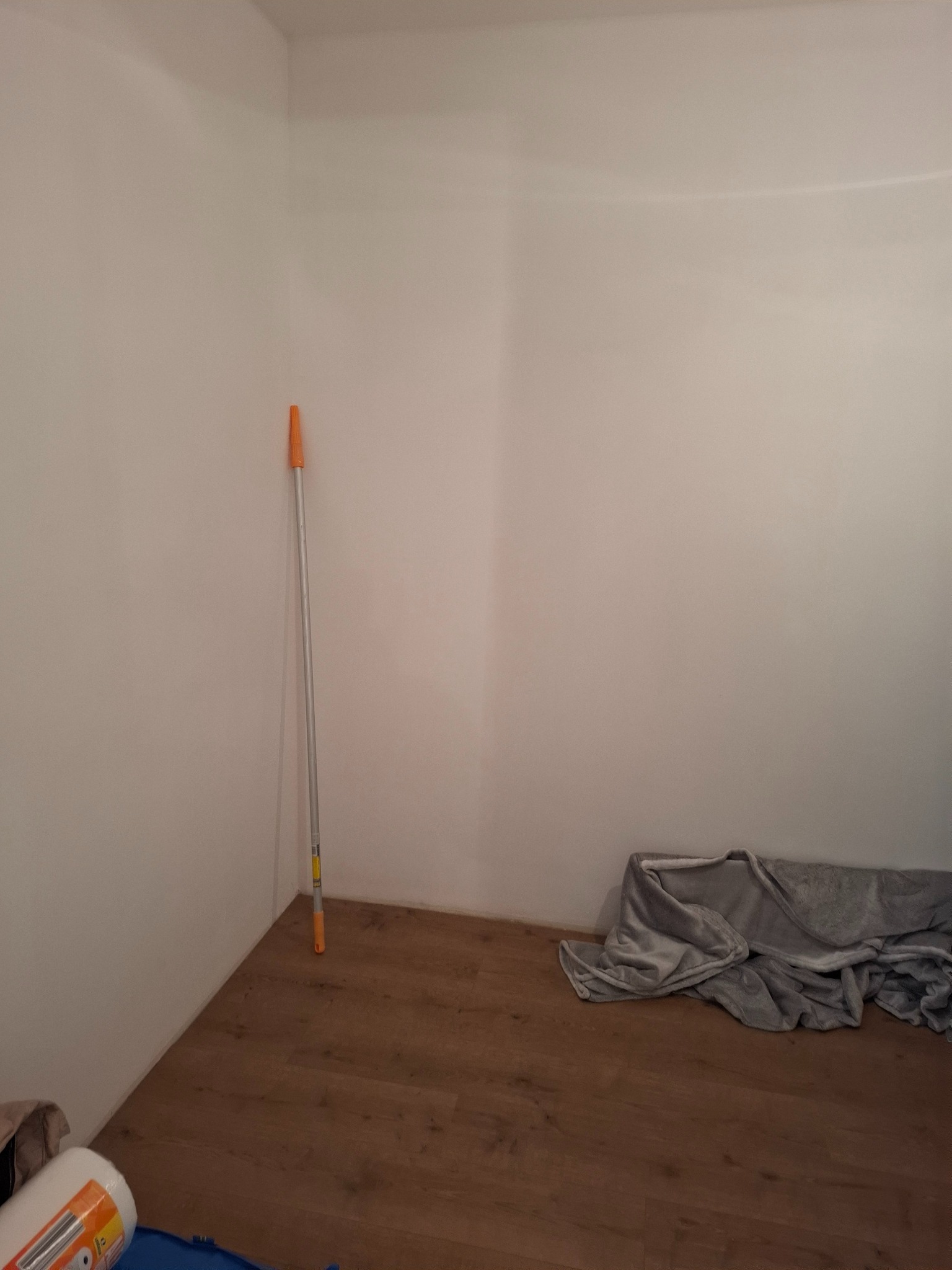Pomalowane na biało ściany w rogu pokoju z drewnianą podłogą. Opiera się o nią kij teleskopowy do malowania. Na podłodze leży szara plandeka i inne akcesoria malarskie.