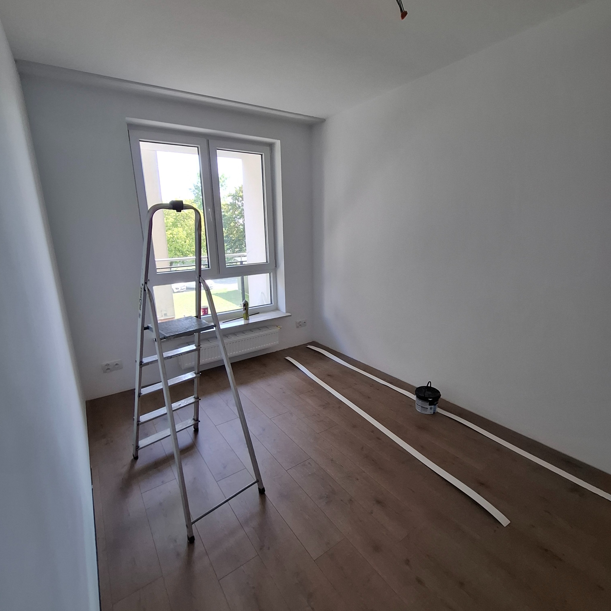 Białe ściany w pokoju z drewnianą podłogą. Widoczna drabina, listwy przypodłogowe i wiadro z farbą. Przygotowanie do malowania.