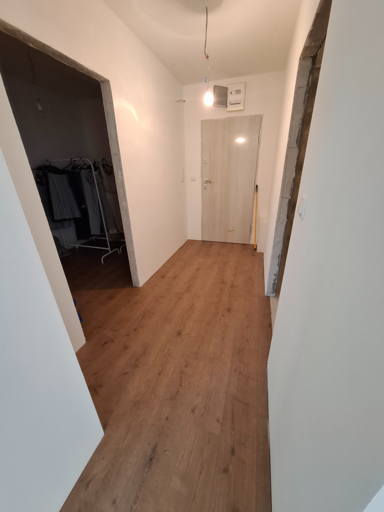 Korytarz w trakcie remontu: świeżo położona podłoga z paneli, surowe ościeżnice drzwiowe, białe ściany. Widoczny wieszak na ubrania w pomieszczeniu obok.