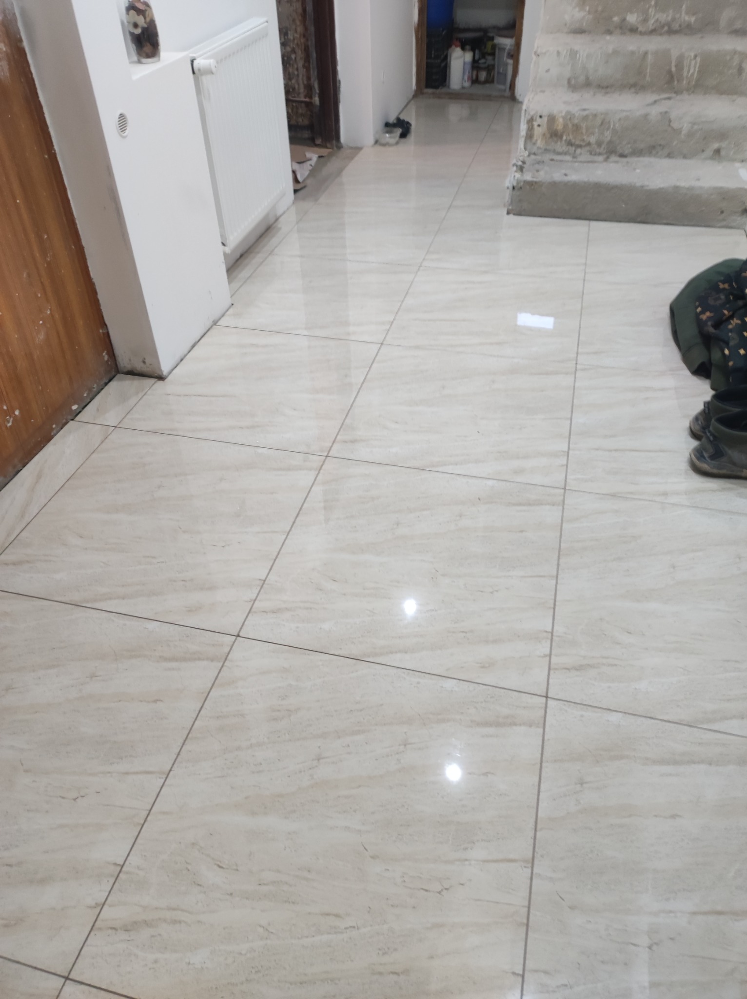 Beżowe, błyszczące płytki podłogowe ułożone w kwadraty, widoczne w korytarzu z grzejnikiem i schodami w tle. Częściowo widoczne buty i ubranie na podłodze.