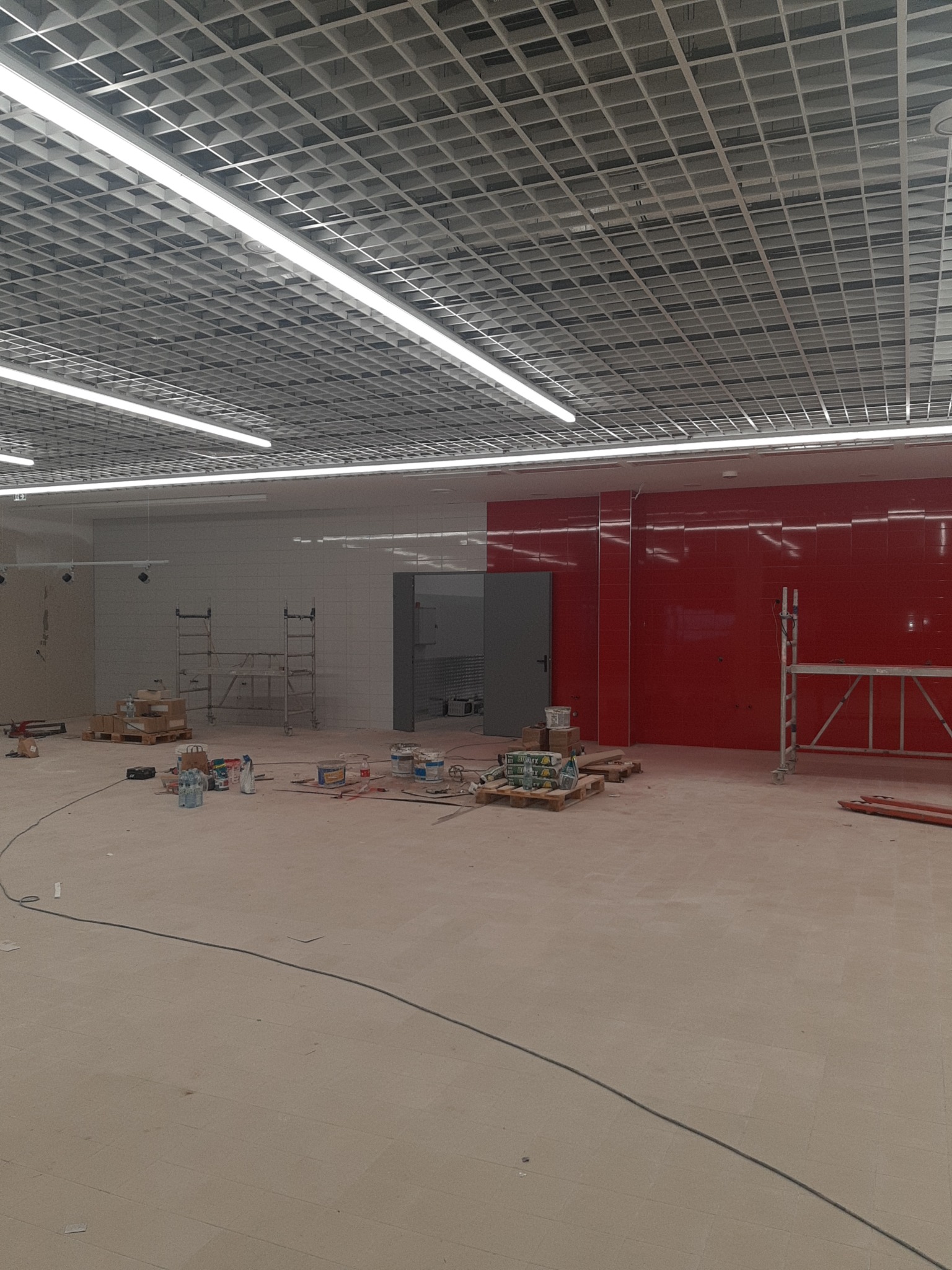 Wnętrze w trakcie prac wykończeniowych: widoczne rusztowania, puszki z farbą i częściowo wykończona ściana z czerwonymi płytkami. Sufit kasetonowy z oświetleniem.