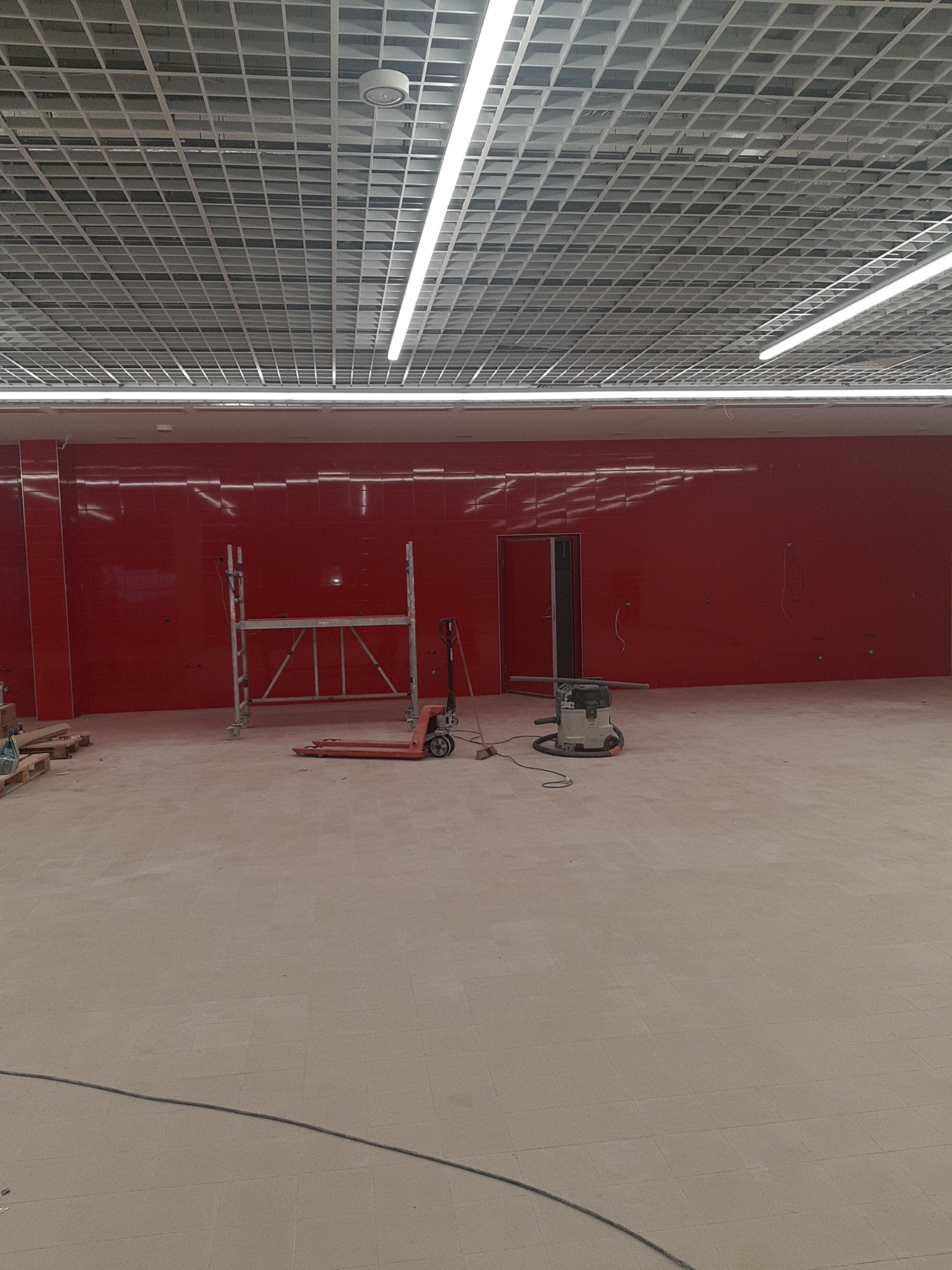 Wnętrze w trakcie prac wykończeniowych: czerwone ściany z płytek, podłoga z terakoty, rusztowanie, wózek paletowy i odkurzacz przemysłowy. Widok na sufit z kratownicą.