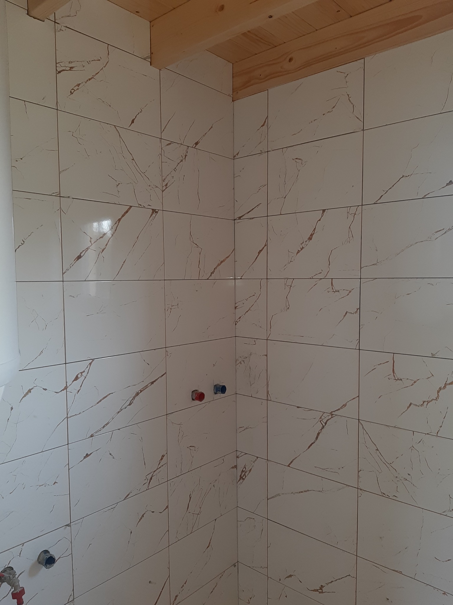 Wykończona ściana w łazience z białych płytek imitujących marmur, widoczne przyłącza wodne i drewniany sufit. Ukończony etap prac glazurniczych.