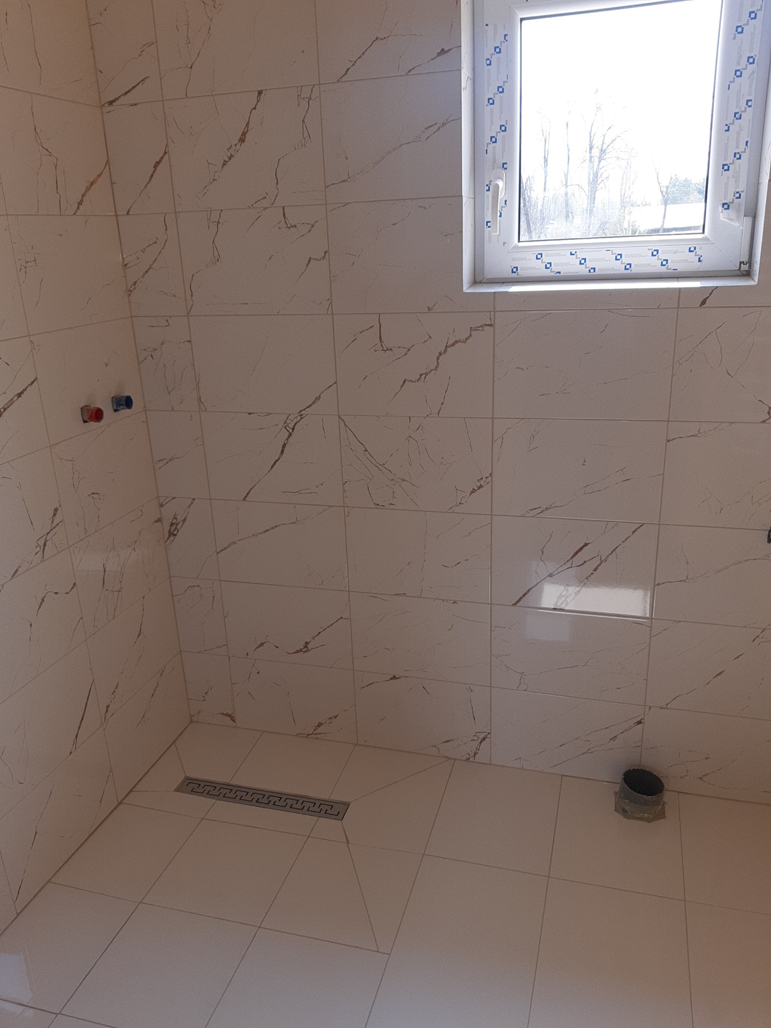 Wykończona łazienka z beżowymi płytkami imitującymi marmur, widoczne okno, odpływ liniowy i przyłącza wodne. Minimalistyczny design.