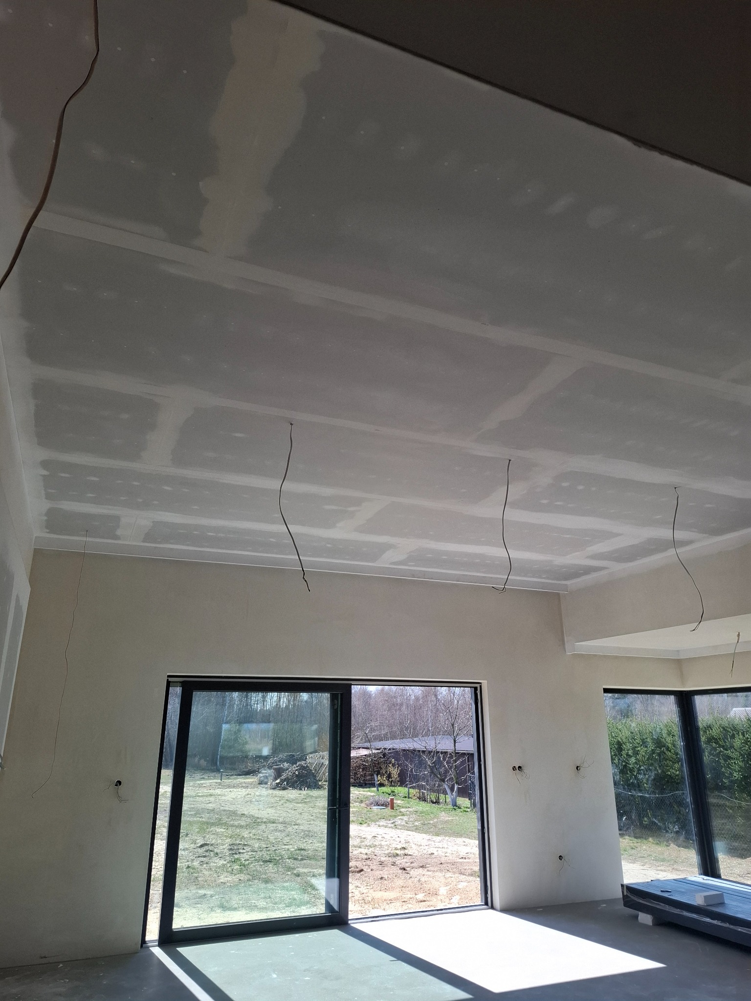 Wnętrze w trakcie szpachlowania: sufit z płyt gipsowo-kartonowych, widoczne łączenia, wystające kable, duże okna z widokiem na zewnątrz. Jasne, naturalne światło.