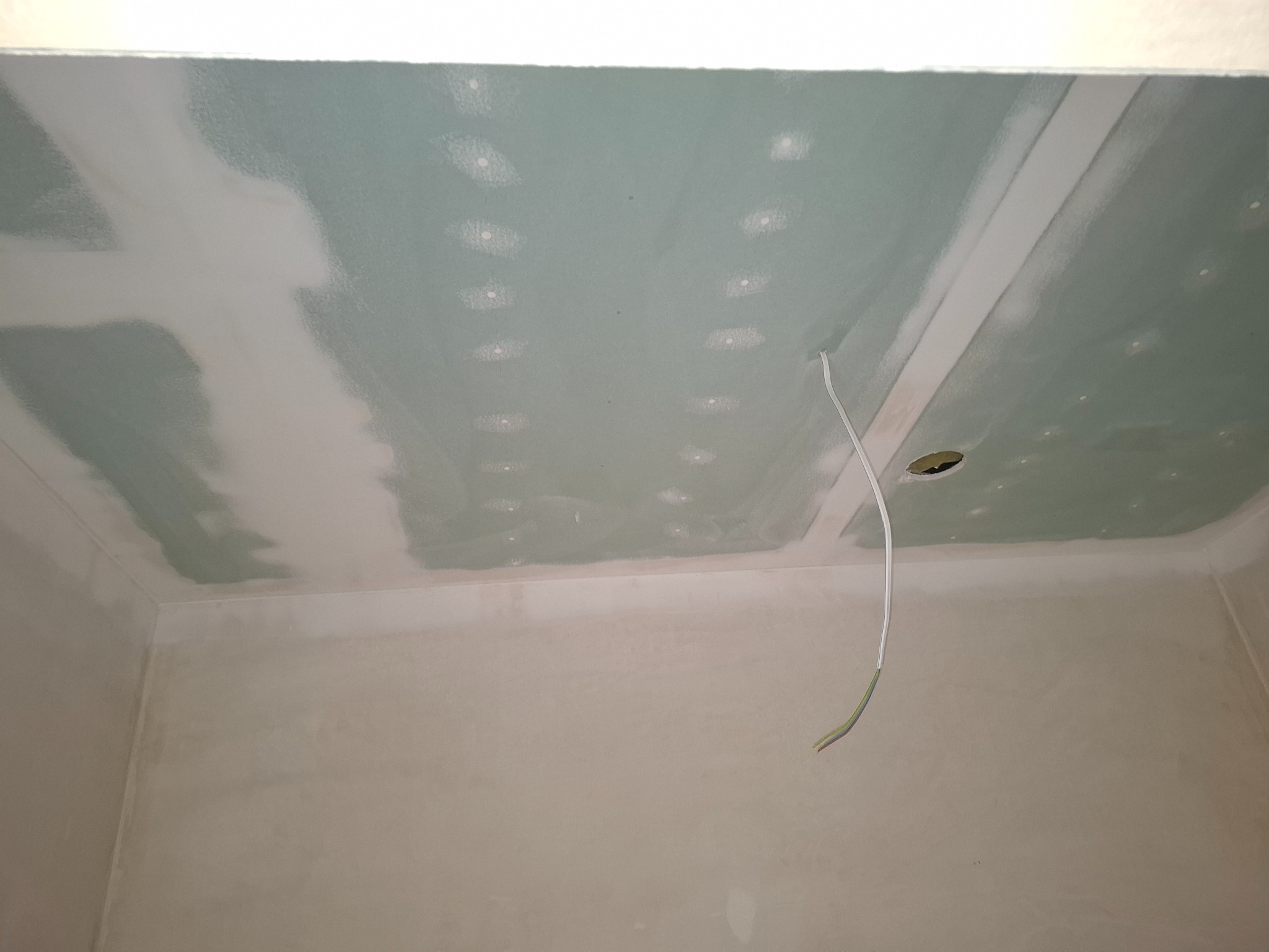 Wnętrze w trakcie szpachlowania: sufit z płyt gipsowo-kartonowych z widocznymi łączeniami i otworem na kabel, ściany po wstępnym szpachlowaniu. Wnętrze w trakcie prac wykończeniowych.