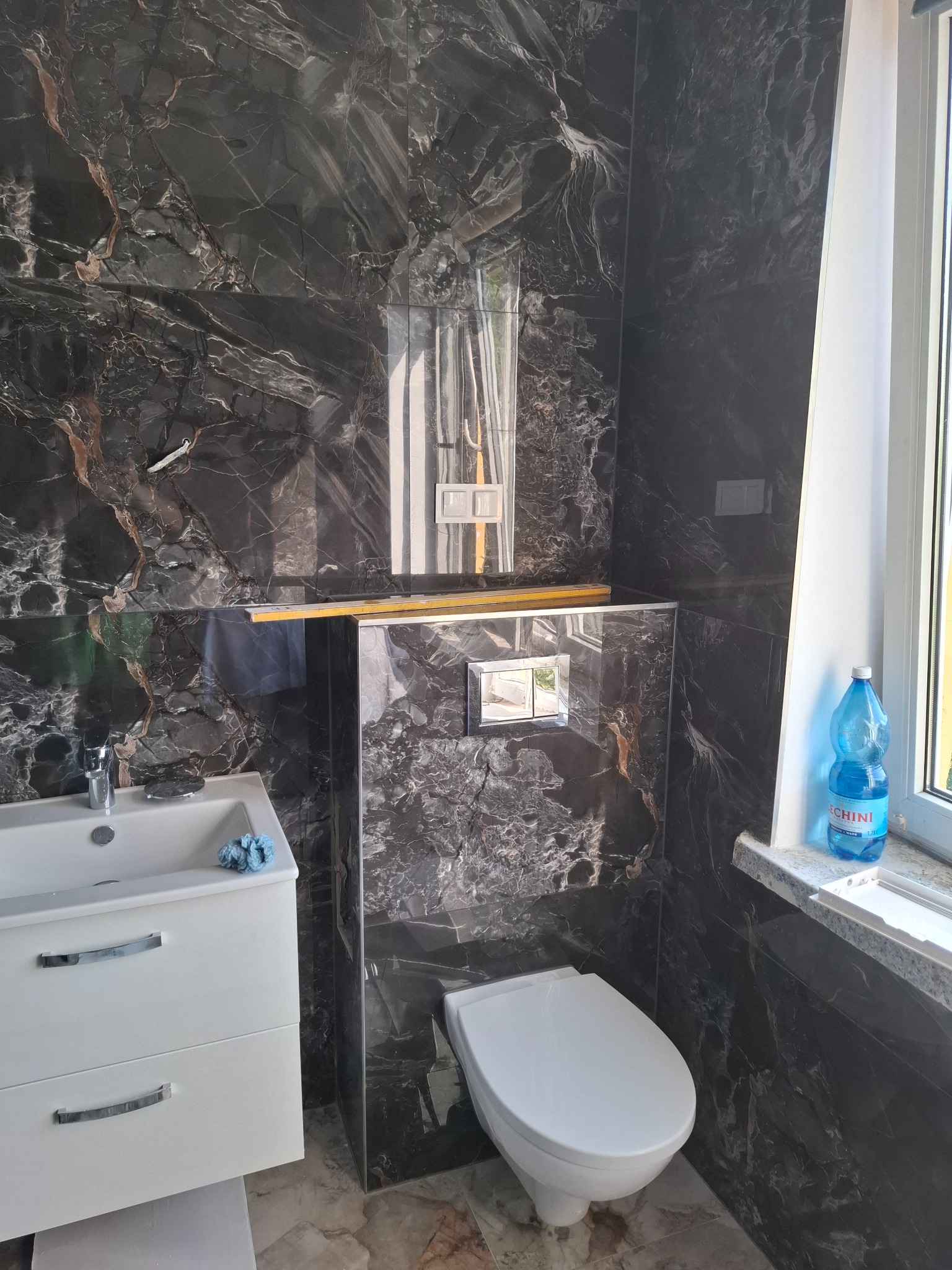 Elegancka łazienka po remoncie: czarne płytki imitujące marmur na ścianach, biała umywalka z szafką i toaleta. Butelka wody na parapecie.
