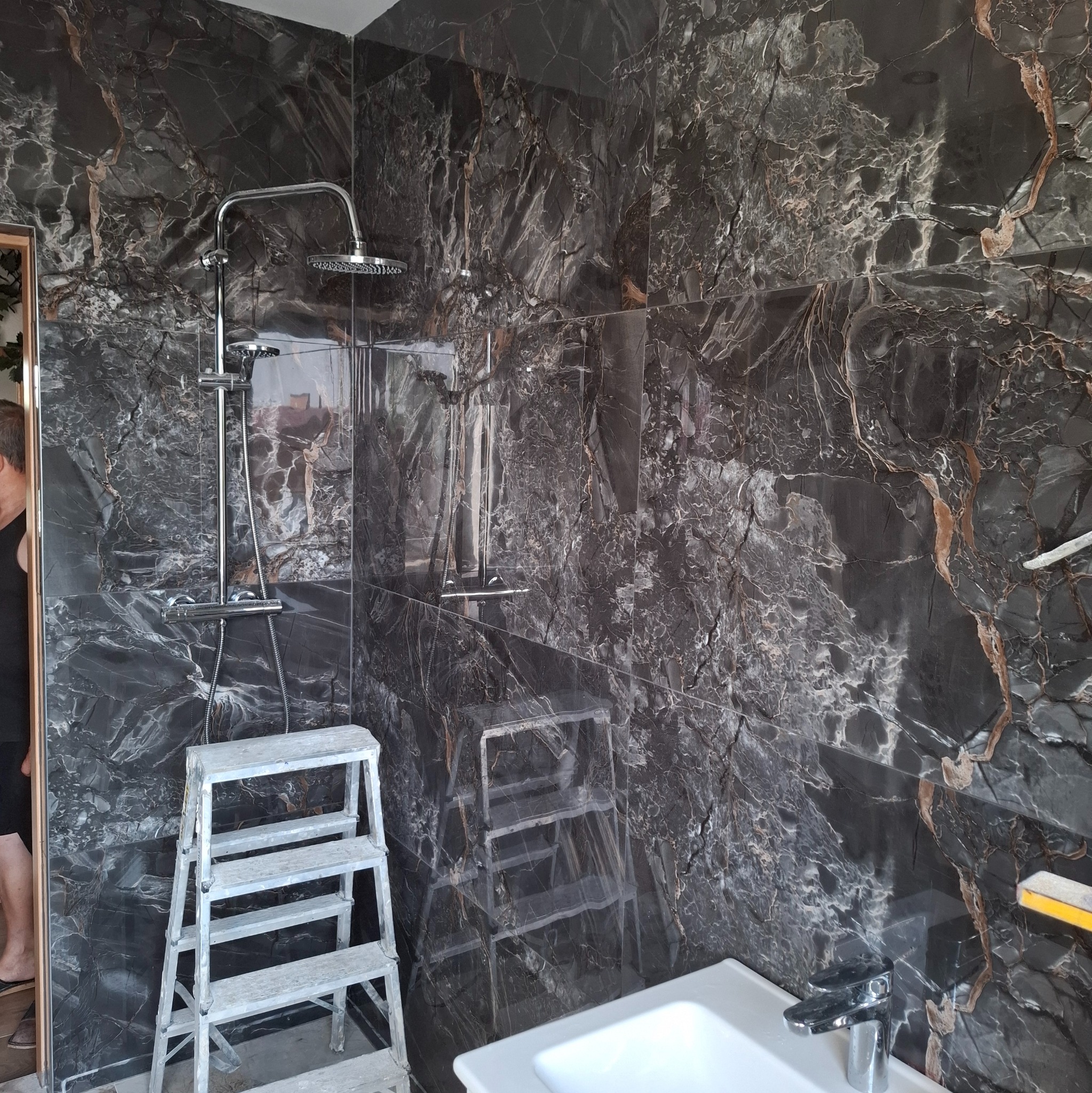 Wykończona łazienka z czarnymi płytkami imitującymi marmur, chromowaną armaturą i drabinką. Widoczny fragment umywalki i drabina glazurnicza.