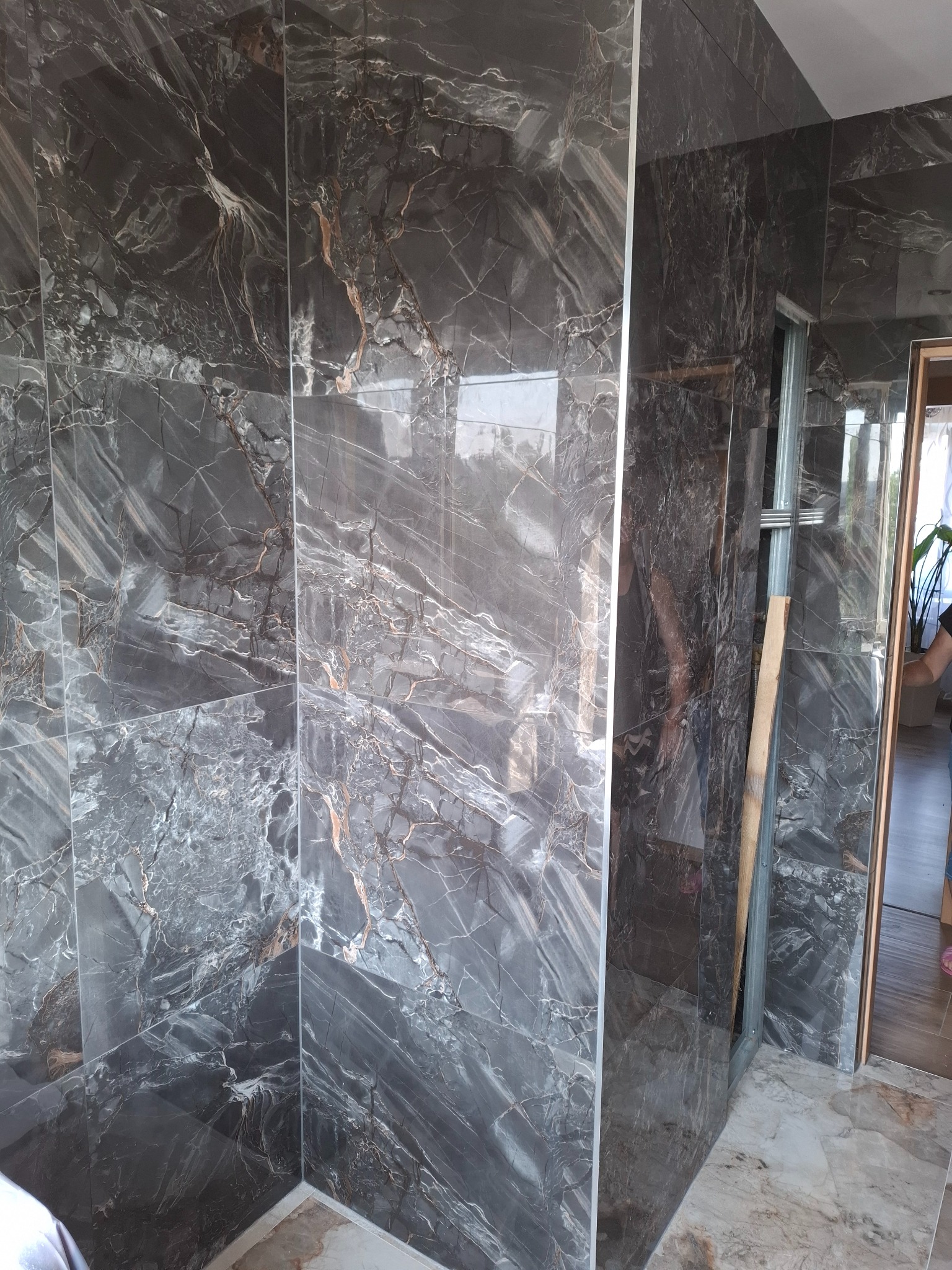 Wykończona ściana z płytek imitujących marmur w odcieniach szarości i brązu, widoczny fragment metalowej konstrukcji i drzwi. Realizacja w trakcie wykańczania.