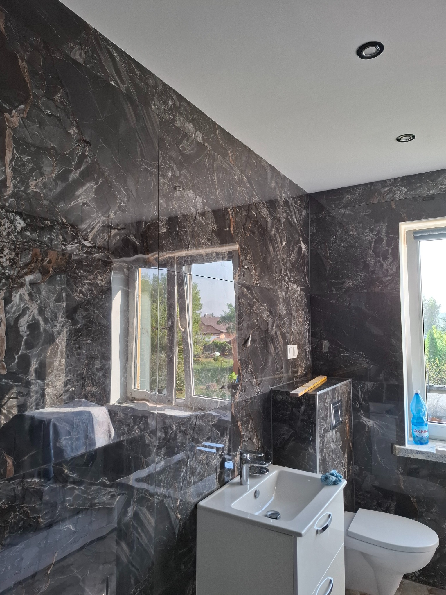 Elegancka łazienka z czarnymi płytkami imitującymi marmur, biała umywalka z szafką i toaleta. Okno z widokiem na zieleń. Nowoczesne oświetlenie sufitowe.