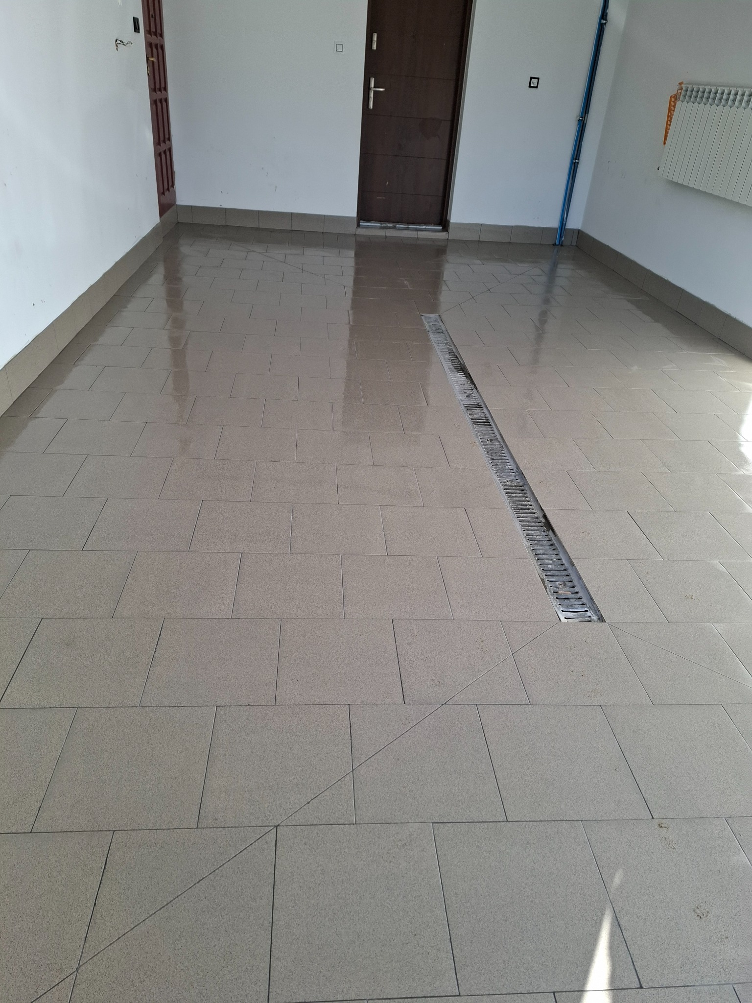 Jasne, kwadratowe płytki podłogowe z odpływem liniowym w nowym pomieszczeniu. Widoczne drzwi wejściowe i grzejnik na ścianie. Świeżo położona podłoga.