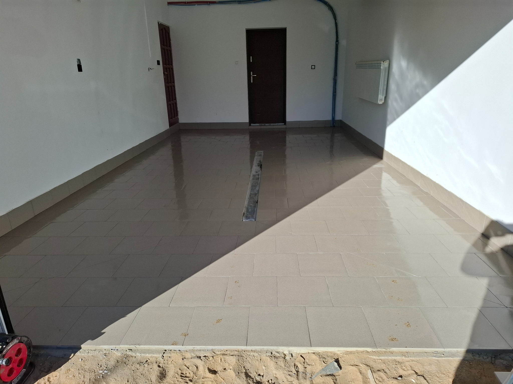 Wyłożona jasnymi płytkami podłoga w pomieszczeniu z białymi ścianami i brązowymi drzwiami. Widoczny odpływ liniowy i cień padający na podłogę.