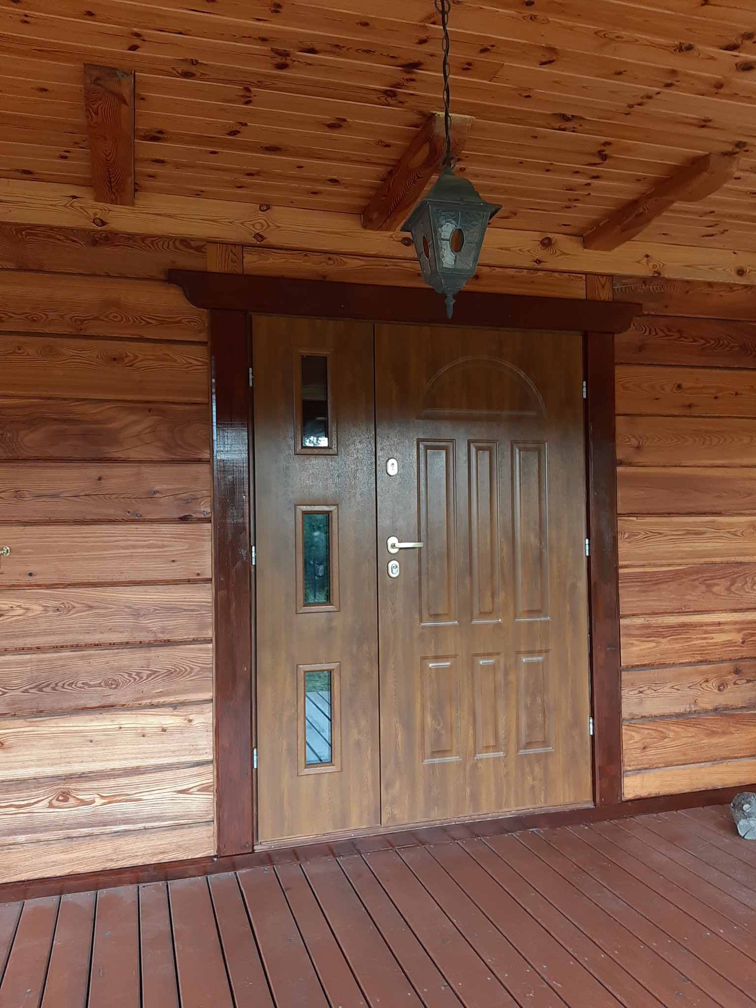 Drewniane drzwi wejściowe z trzema szybkami, zamontowane w domu z bali, oświetlone wiszącą lampą w stylu retro. Widoczny fragment drewnianego tarasu.