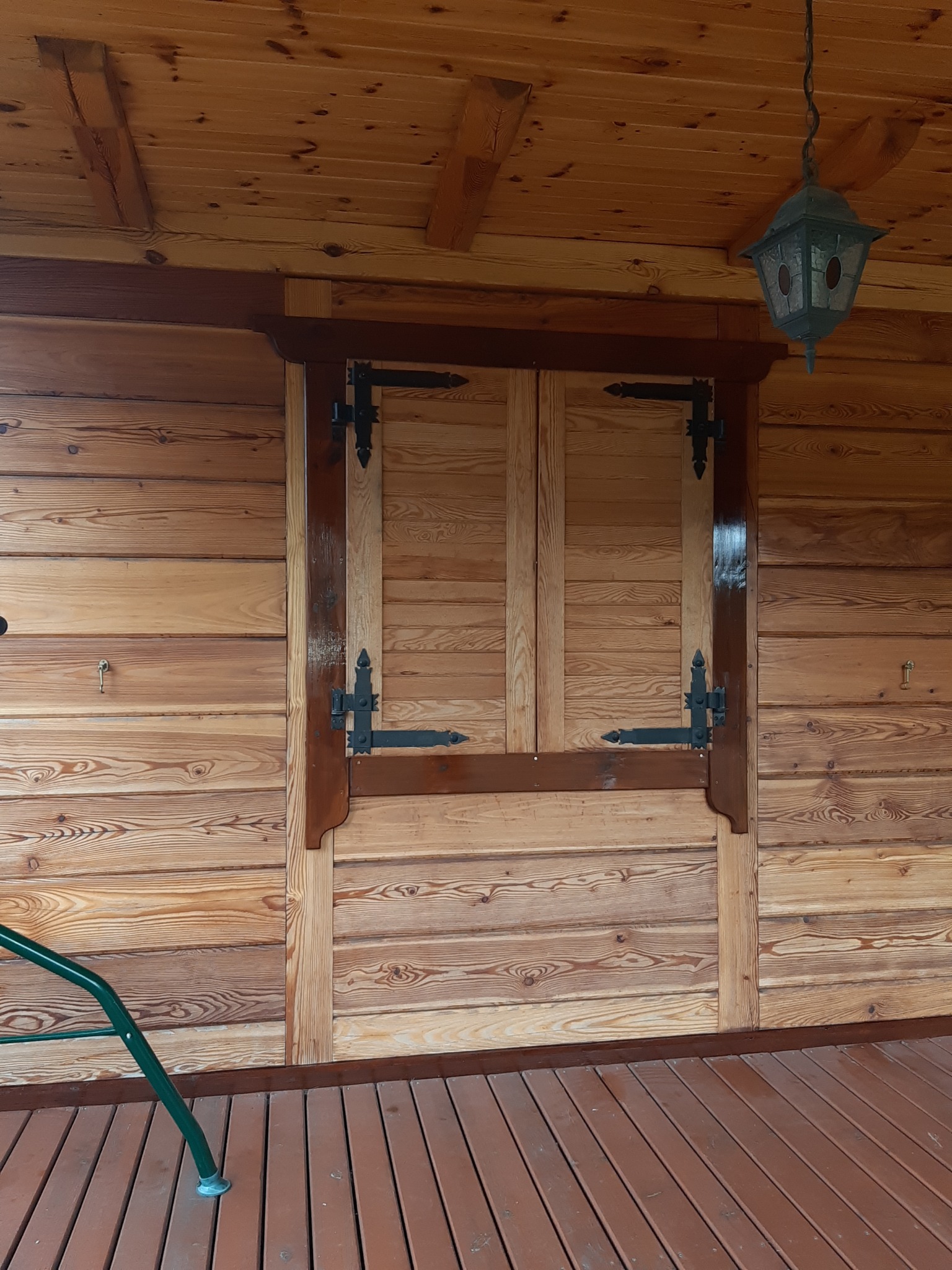 Drewniana ściana z zamkniętymi okiennicami z ozdobnymi zawiasami i wiszącą lampą. Widoczny fragment drewnianej podłogi i element huśtawki.