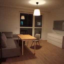 RM-BUD wykończenie wnętrz mieszkań i domów - Jasne wnętrze pokoju dziennego z szarą sofą, drewnianym stołem i komodą. Na ścianie zabezpieczony folią telewizor. Parkiet na podłodze. Dwa okna balkonowe.