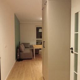 RM-BUD wykończenie wnętrz mieszkań i domów - Jasne wnętrze mieszkania z drewnianą podłogą, szarą sofą i szafą w zabudowie. Widok z korytarza na salon, drzwi z przeszkleniami po prawej.