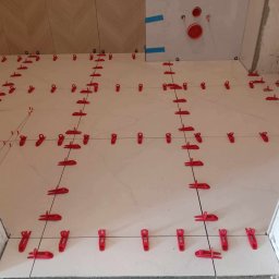 RM-BUD wykończenie wnętrz mieszkań i domów - Jasne płytki podłogowe z czerwonymi klipsami poziomującymi w trakcie układania. Widoczne fragmenty ścian z instalacją. Ułożony wzór na ścianie w tle.