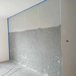 RM-BUD wykończenie wnętrz mieszkań i domów - Ściana z płyt kartonowo-gipsowych w trakcie szpachlowania, zabezpieczona niebieską taśmą malarską. Widoczne gniazdka elektryczne i surowy beton na podłodze.