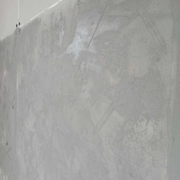 RM-BUD wykończenie wnętrz mieszkań i domów - Świeżo nałożona, szara gładź szpachlowa na ścianie, widoczne ślady po szpachelce. Ujęcie z dołu, podkreślające fakturę i nierówności. Oświetlenie naturalne, dzienne.