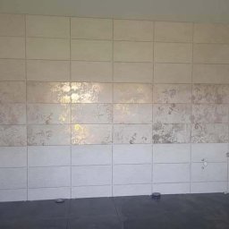 RM-BUD wykończenie wnętrz mieszkań i domów - Ściana wyłożona beżowymi płytkami z delikatnym wzorem, częściowo ułożona, widoczne otwory na instalacje, ciemna podłoga, surowe wnętrze.