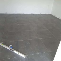 RM-BUD wykończenie wnętrz mieszkań i domów - Układanie ciemnoszarych płytek podłogowych w pomieszczeniu, z użyciem poziomicy i elementów dystansowych, widoczne fugi i fragment ściany.