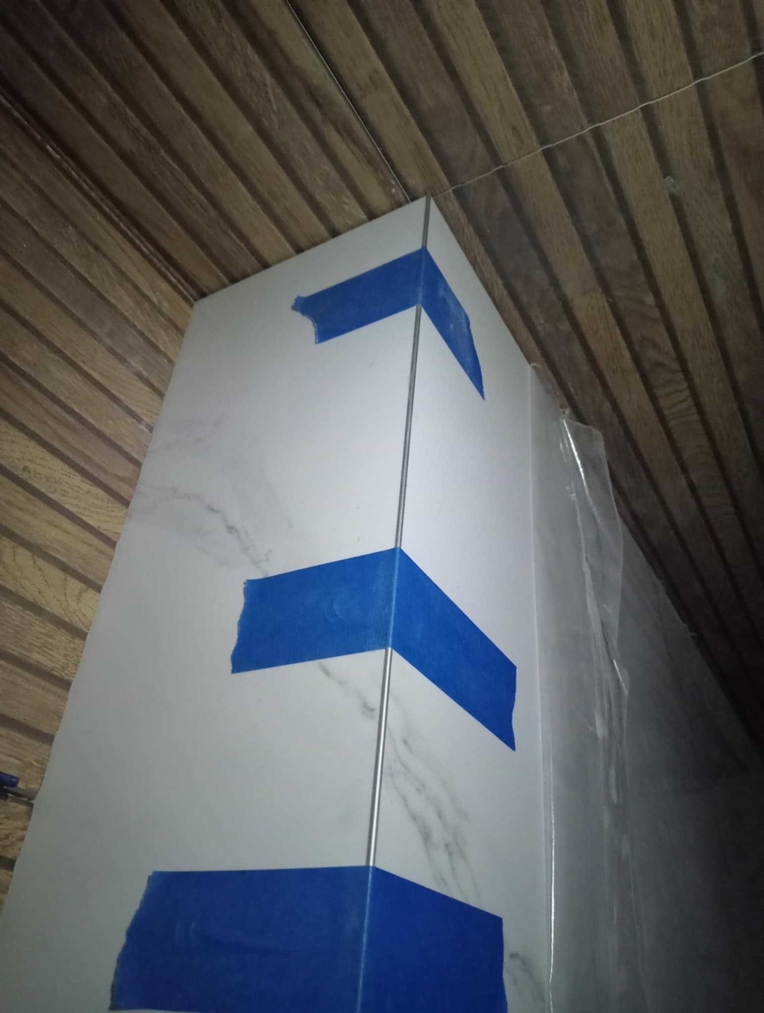 Wykończony narożnik z białych płytek imitujących marmur, zabezpieczony niebieską taśmą malarską. Widoczna listwa wykończeniowa i drewniana boazeria na suficie.