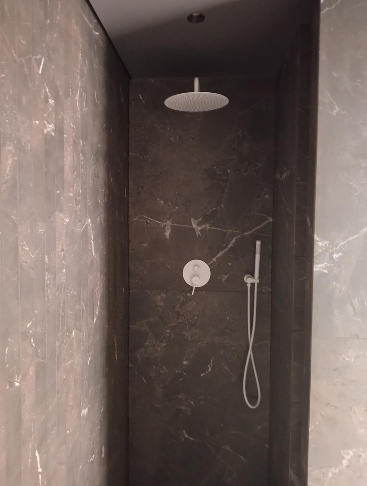 Nowoczesna kabina prysznicowa z ciemnymi płytkami imitującymi marmur, deszczownicą sufitową i minimalistyczną armaturą w odcieniach bieli. Eleganckie wykończenie.