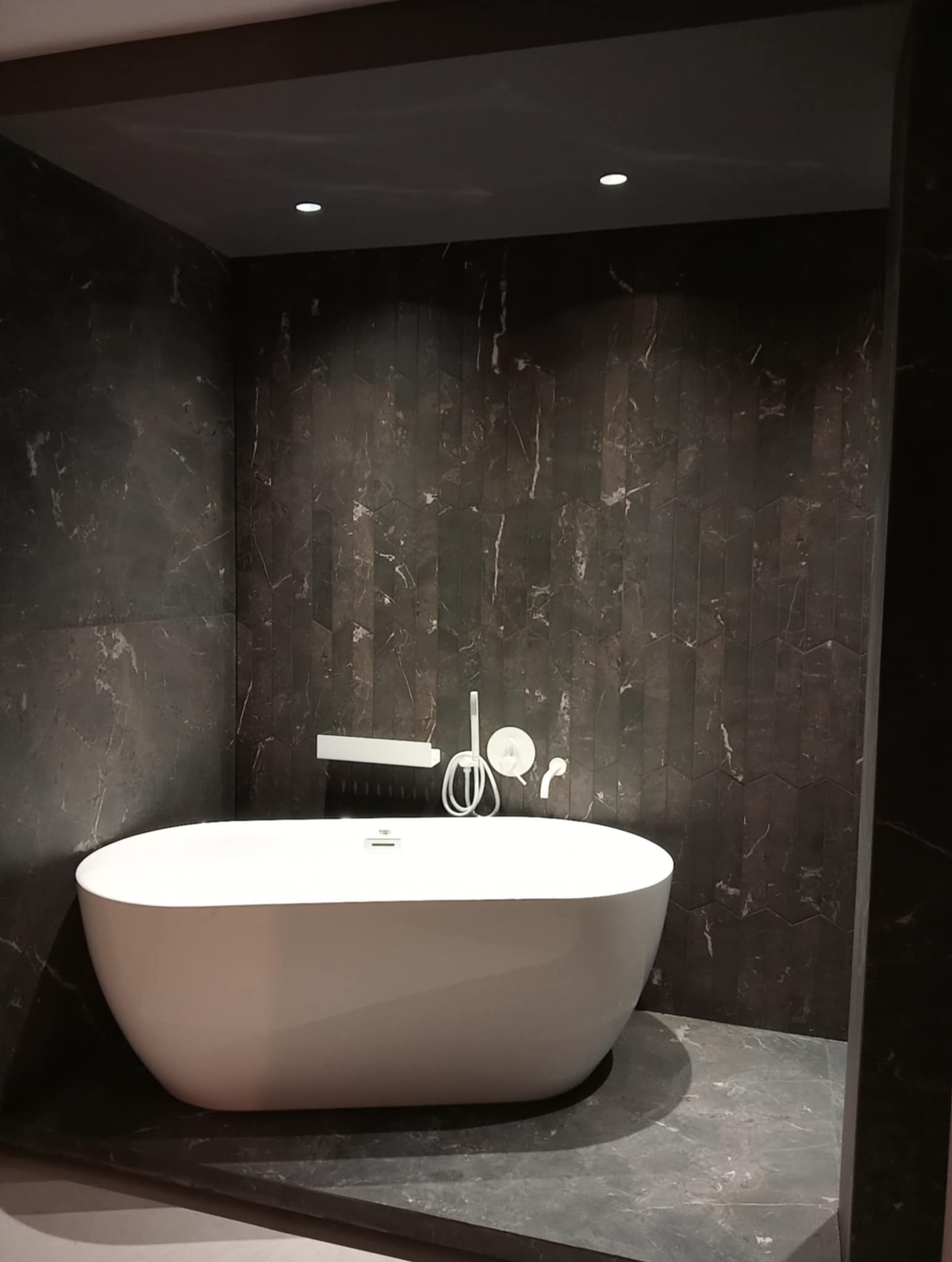 Nowoczesna łazienka z wolnostojącą białą wanną na tle ściany wyłożonej ciemnym marmurem w jodełkę. Minimalistyczna armatura i punktowe oświetlenie.