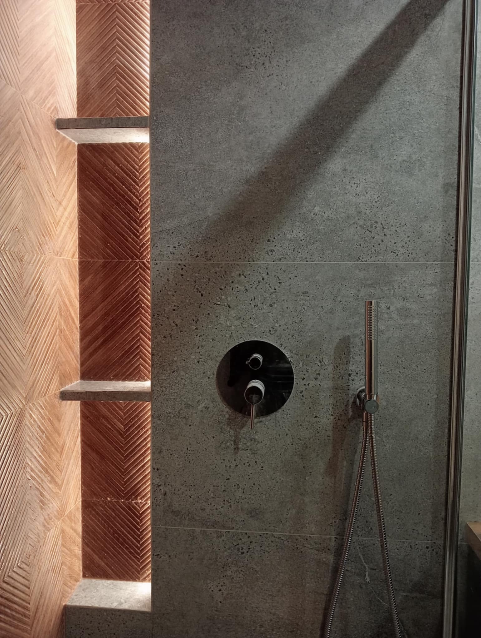 Nowoczesna łazienka: szara ściana z armaturą i dekoracyjna wnęka z drewnopodobnymi płytkami ułożonymi we wzór jodełki, oświetlona od góry.