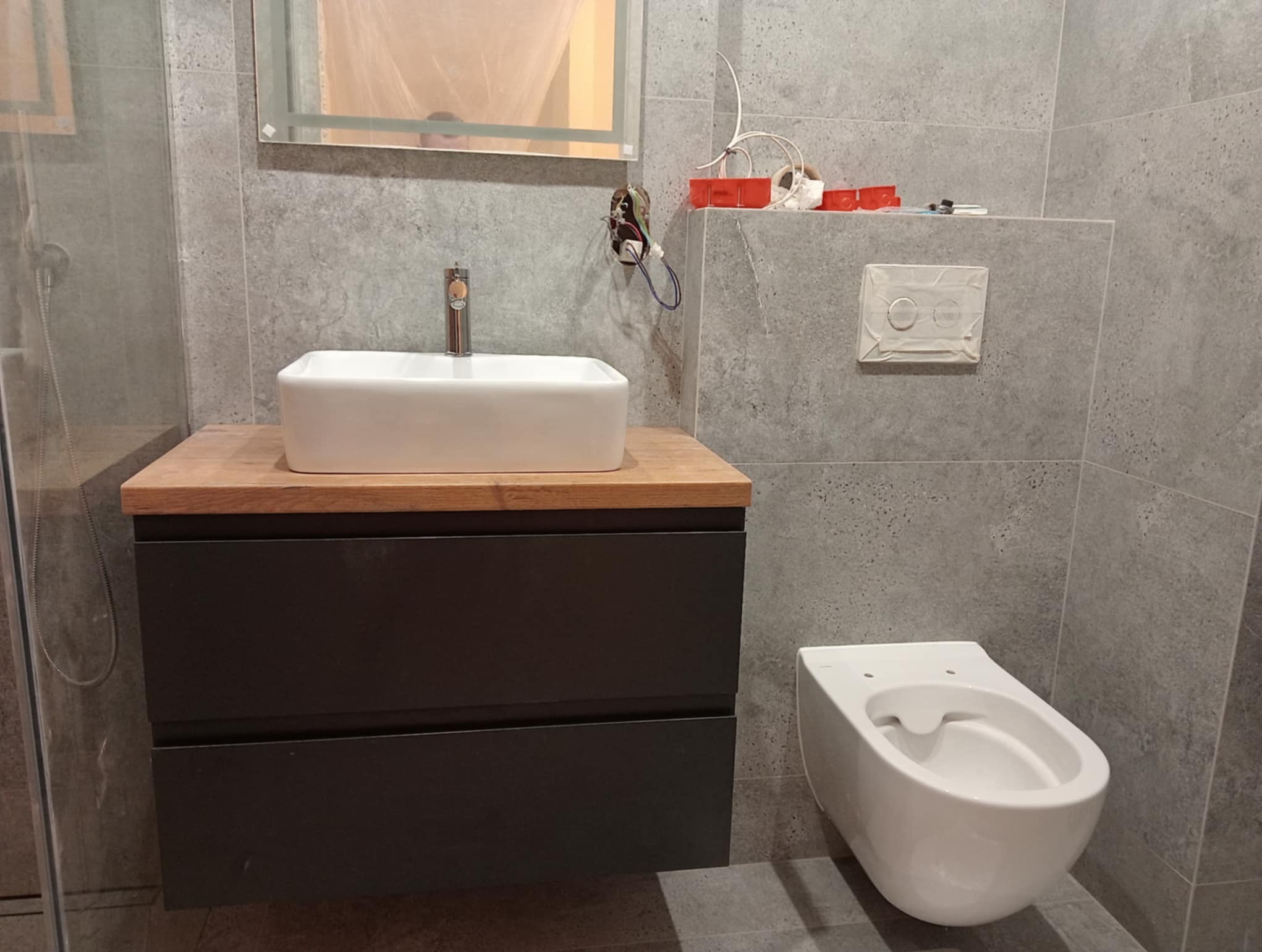 Szara łazienka po remoncie: podwieszana toaleta, szafka z umywalką na drewnianym blacie, szklana kabina prysznicowa. Widoczne niedokończone prace elektryczne.
