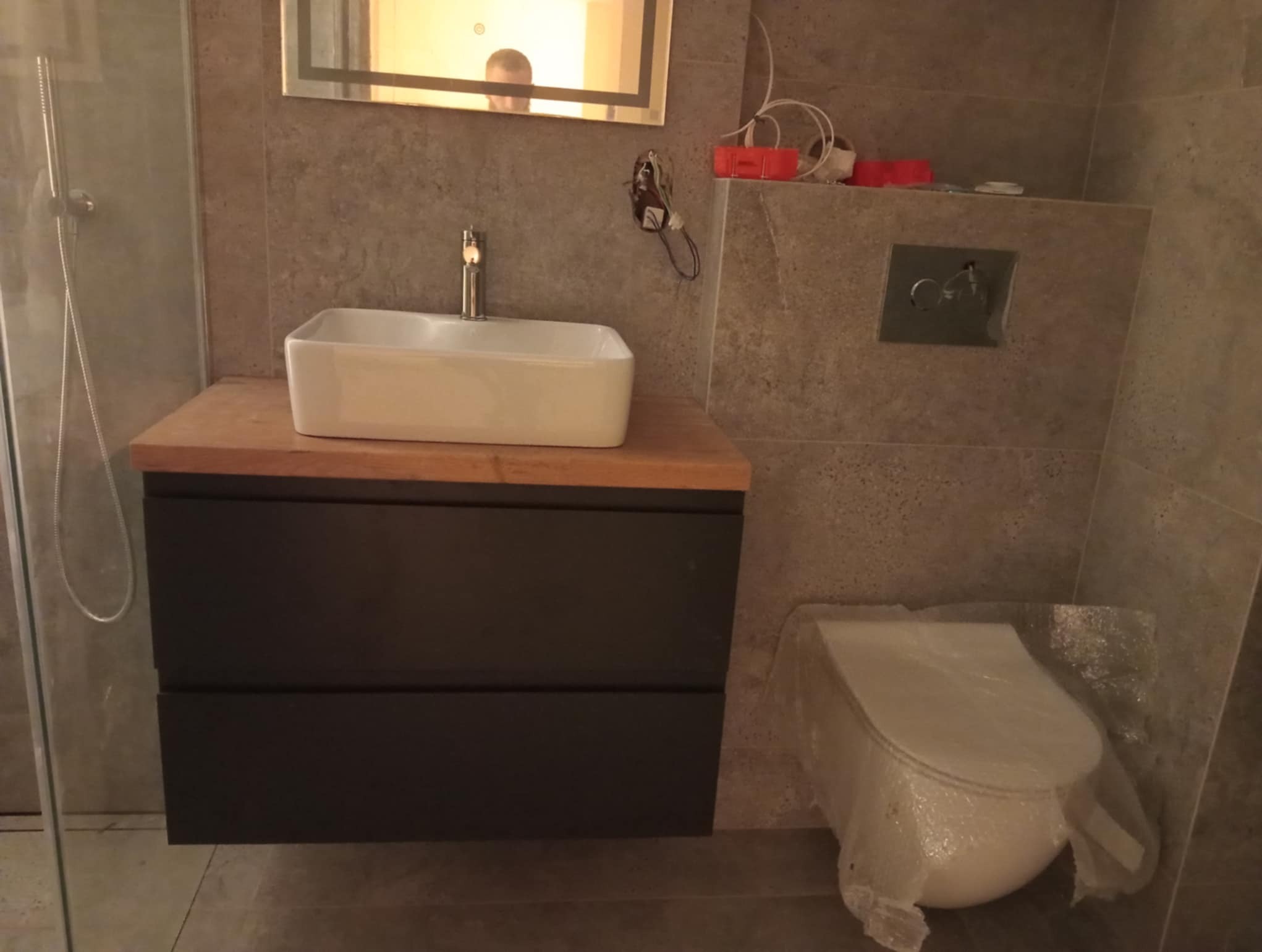 Nowoczesna łazienka z umywalką nablatową na drewnianym blacie i szarą armaturą. Toaleta zabezpieczona folią, widoczne płytki imitujące kamień i fragment kabiny prysznicowej.