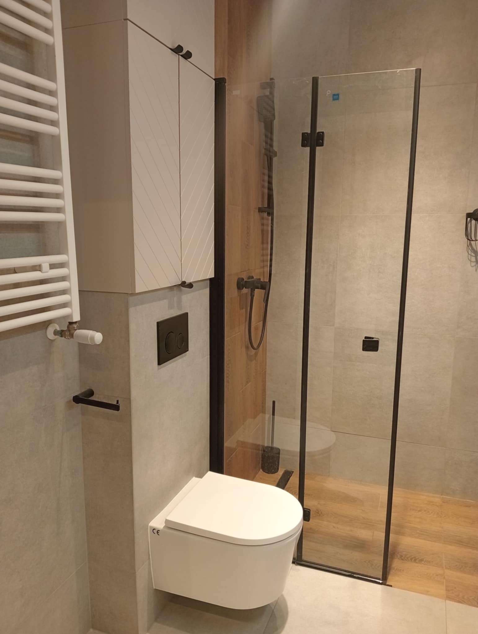 Nowoczesna łazienka z kabiną prysznicową typu walk-in, podwieszaną toaletą i grzejnikiem drabinkowym. Uwagę zwraca szafka z geometrycznym wzorem i czarne akcenty.