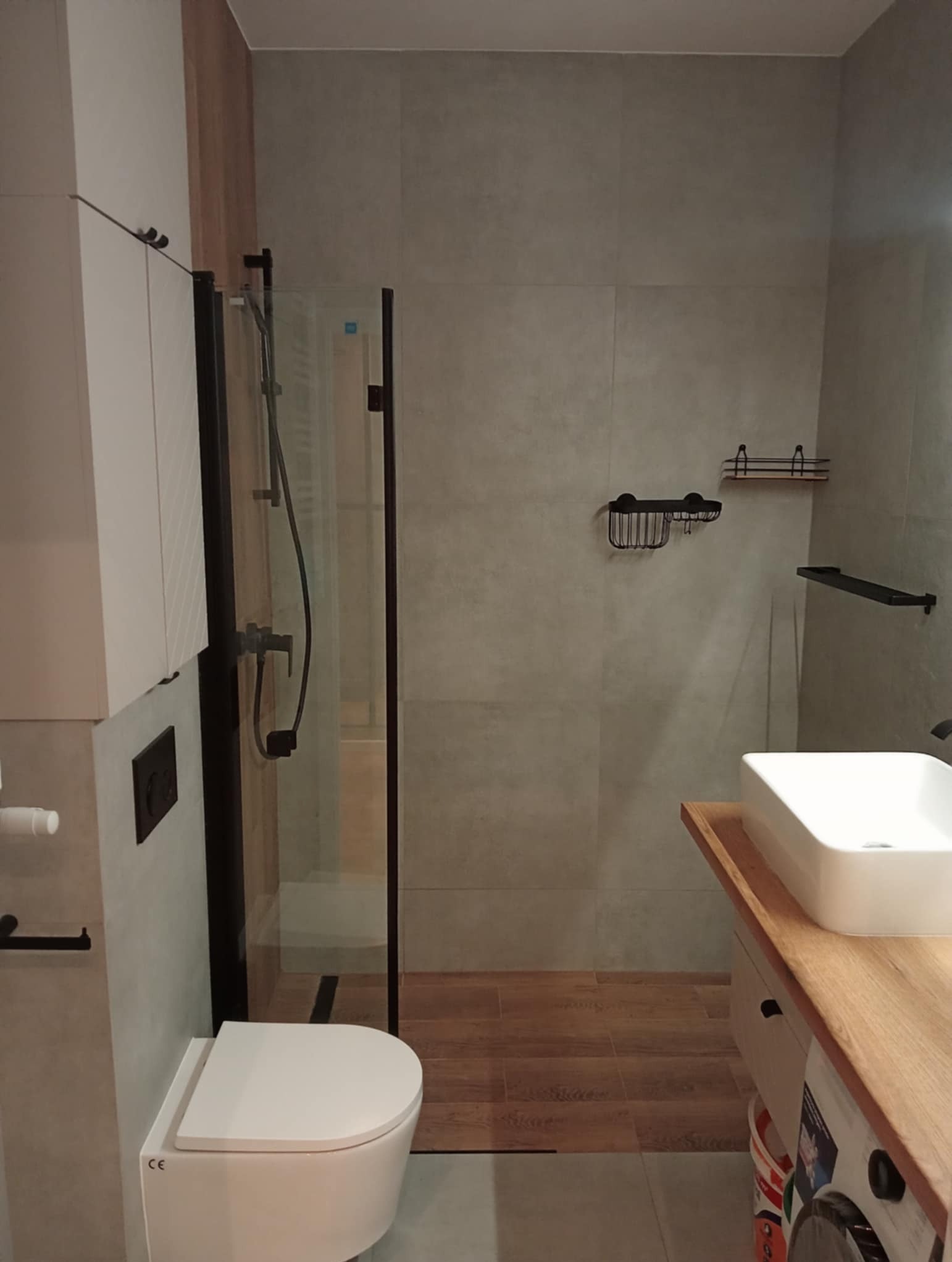 Nowoczesna łazienka z kabiną prysznicową typu walk-in, umywalką nablatową na drewnianym blacie i podwieszaną toaletą. Szare płytki i drewnopodobna podłoga.