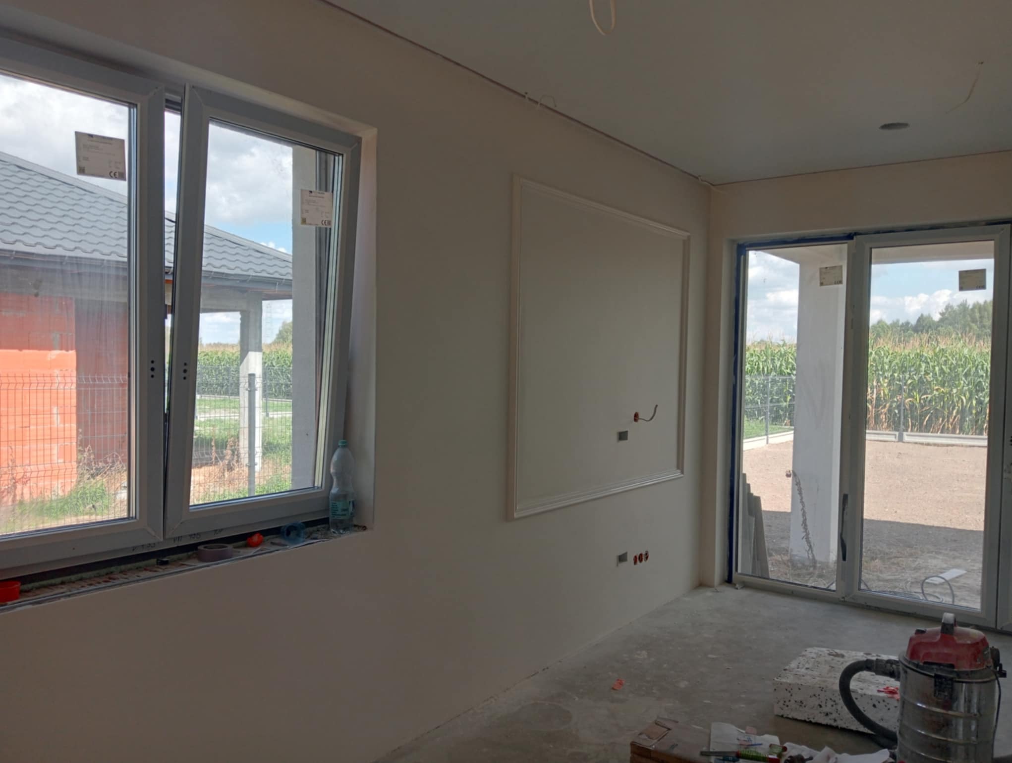 Wnętrze w trakcie wykańczania: białe ściany z dekoracyjną ramą, okno z widokiem na dom i pole kukurydzy, przesuwne drzwi balkonowe, narzędzia na podłodze.