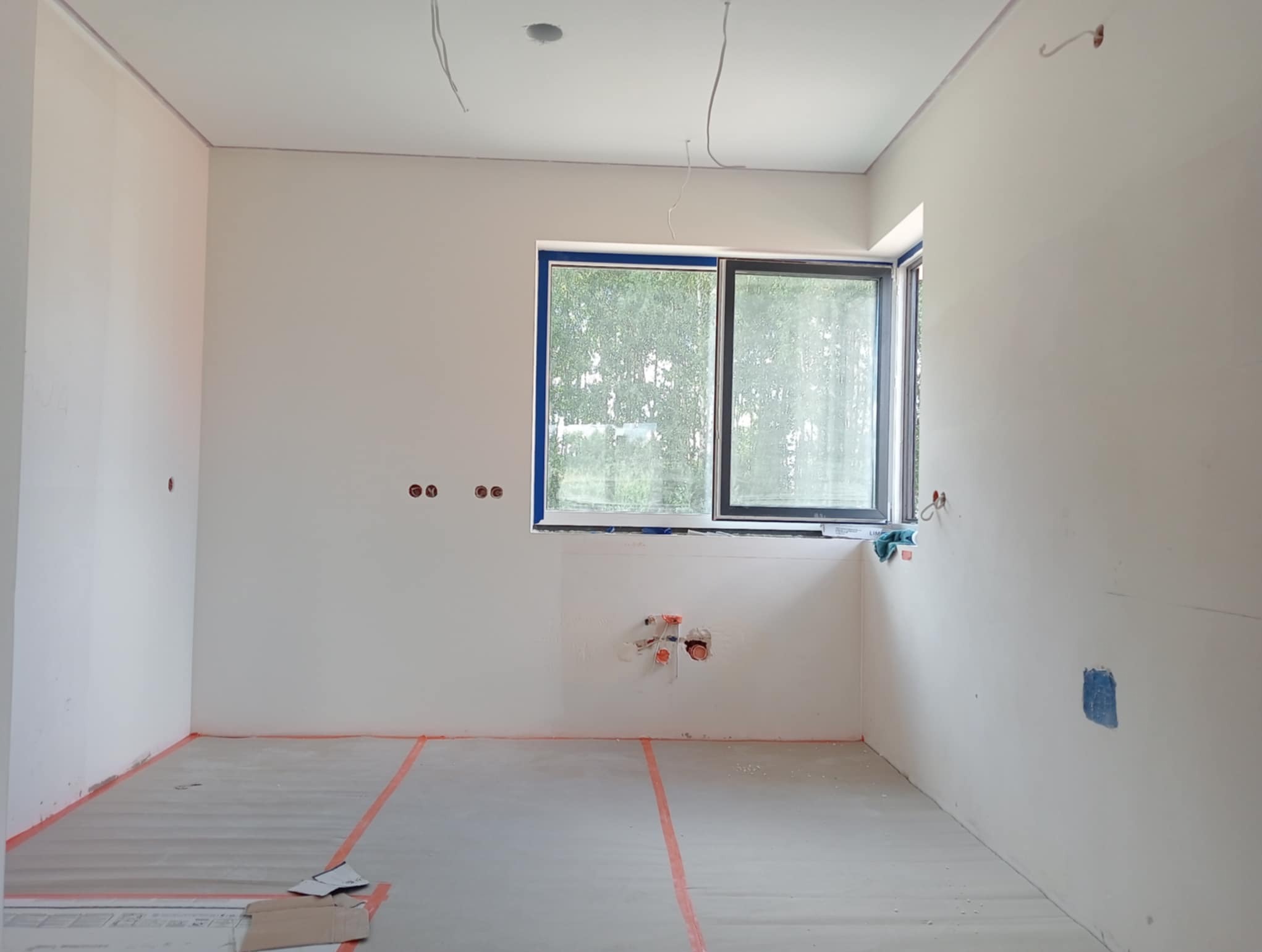 Wnętrze w trakcie prac wykończeniowych: ściany przygotowane do malowania, widoczne instalacje elektryczne i hydrauliczne, okno z niebieską taśmą zabezpieczającą.
