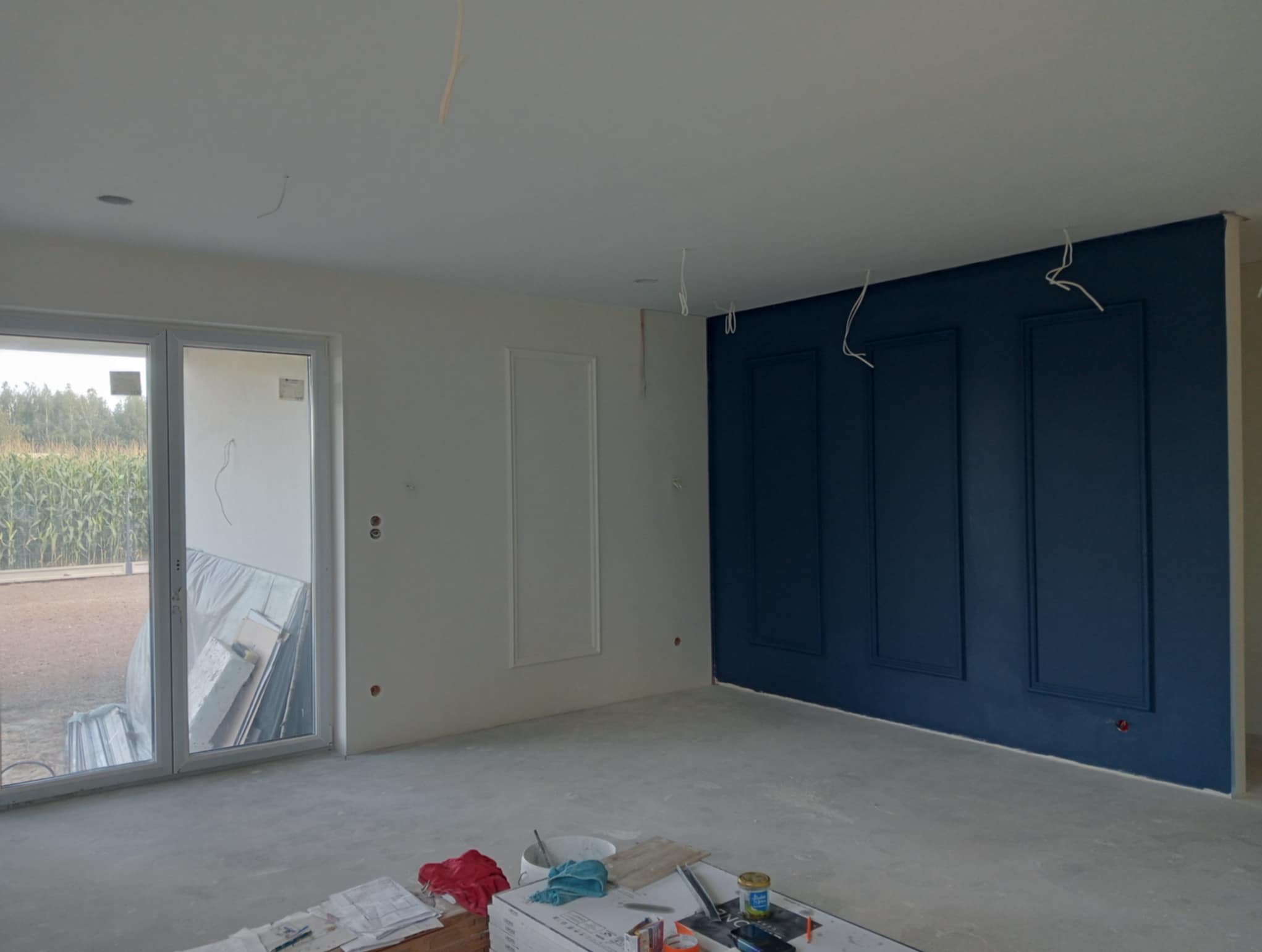 Wnętrze w trakcie wykańczania: białe ściany z dekoracyjnymi listwami, granatowa ściana z panelami, surowa podłoga, okno z widokiem na zewnątrz. Widoczne narzędzia i materiały.