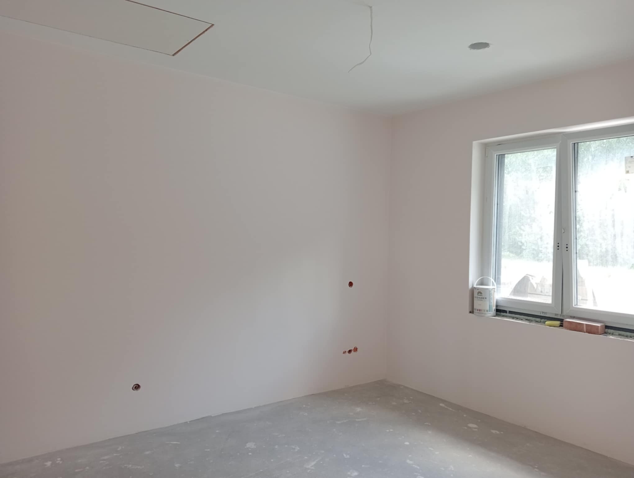 Jasny pokój po malowaniu ścian na delikatny róż, widoczne puszki instalacyjne, okno z widokiem na zieleń, puszka farby na parapecie. Podłoga szara, surowa.
