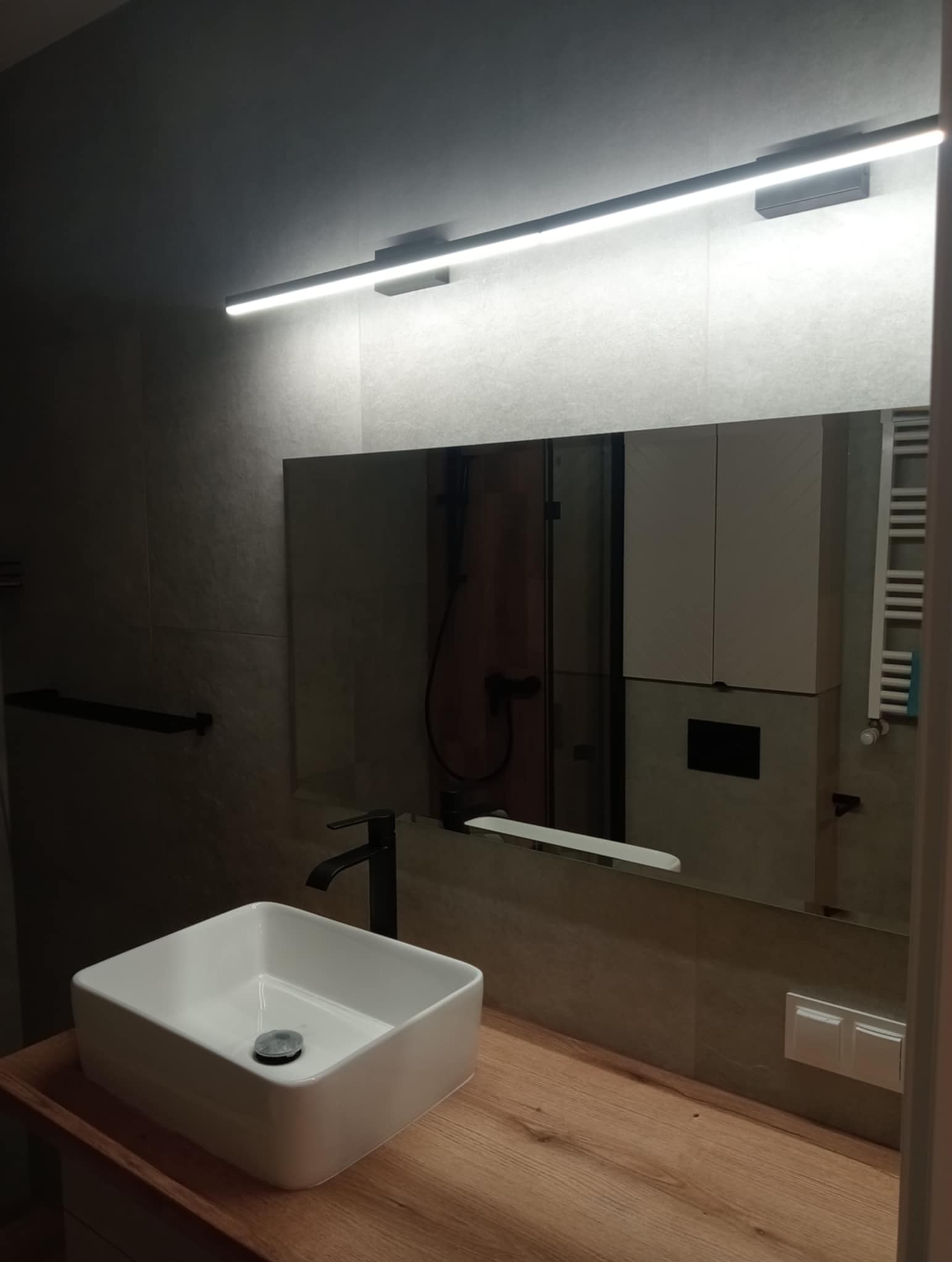 Nowoczesna łazienka z kwadratową umywalką nablatową, czarną armaturą i podświetlanym lustrem. Minimalistyczny design z drewnianym blatem i szarą płytką.