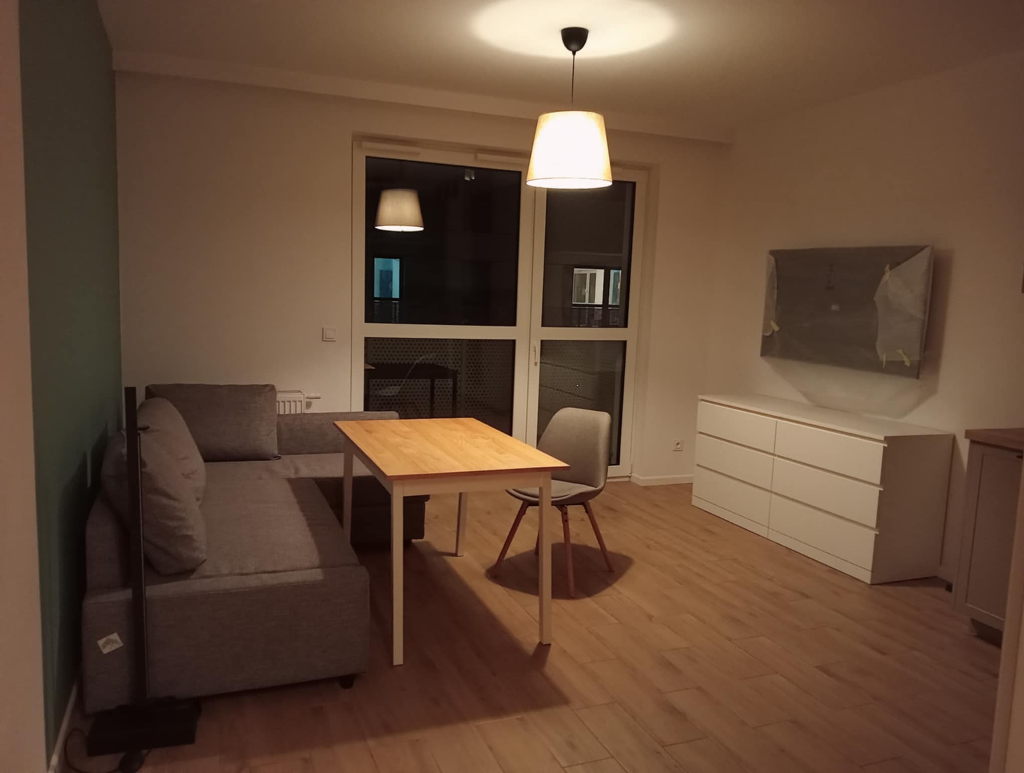 Jasne wnętrze pokoju dziennego z szarą sofą, drewnianym stołem i komodą. Na ścianie zabezpieczony folią telewizor. Parkiet na podłodze. Dwa okna balkonowe.