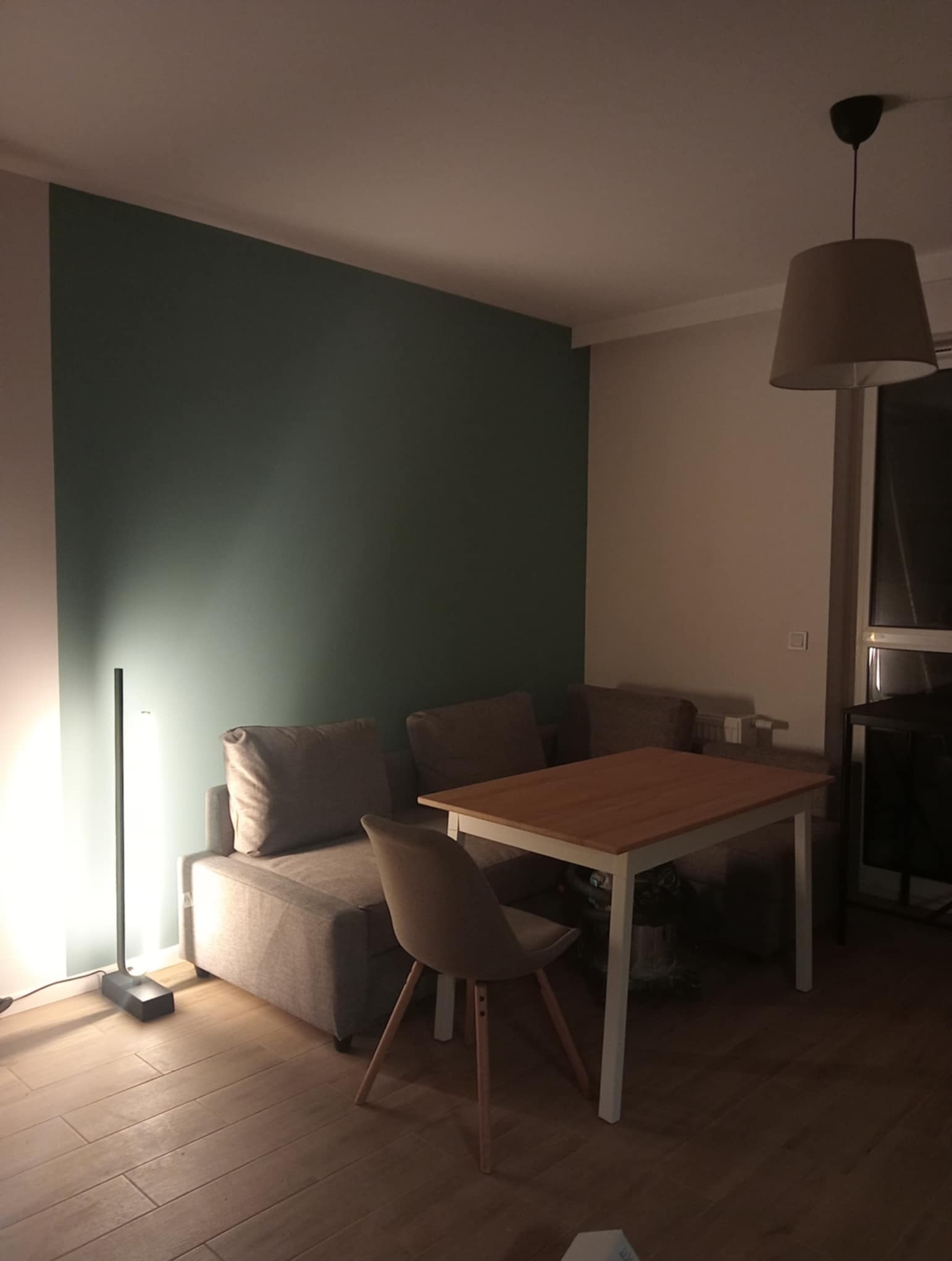Salon po malowaniu: ściana w odcieniach zieleni, beżowa sofa, drewniany stół z białymi nogami, lampa podłogowa i wisząca. Minimalistyczny wystrój wnętrza.